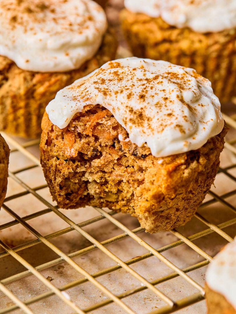 Healthy carrot cake muffins with greek yogurt frosting on a wire cooling rack. The one in front has a bite taken out of it.