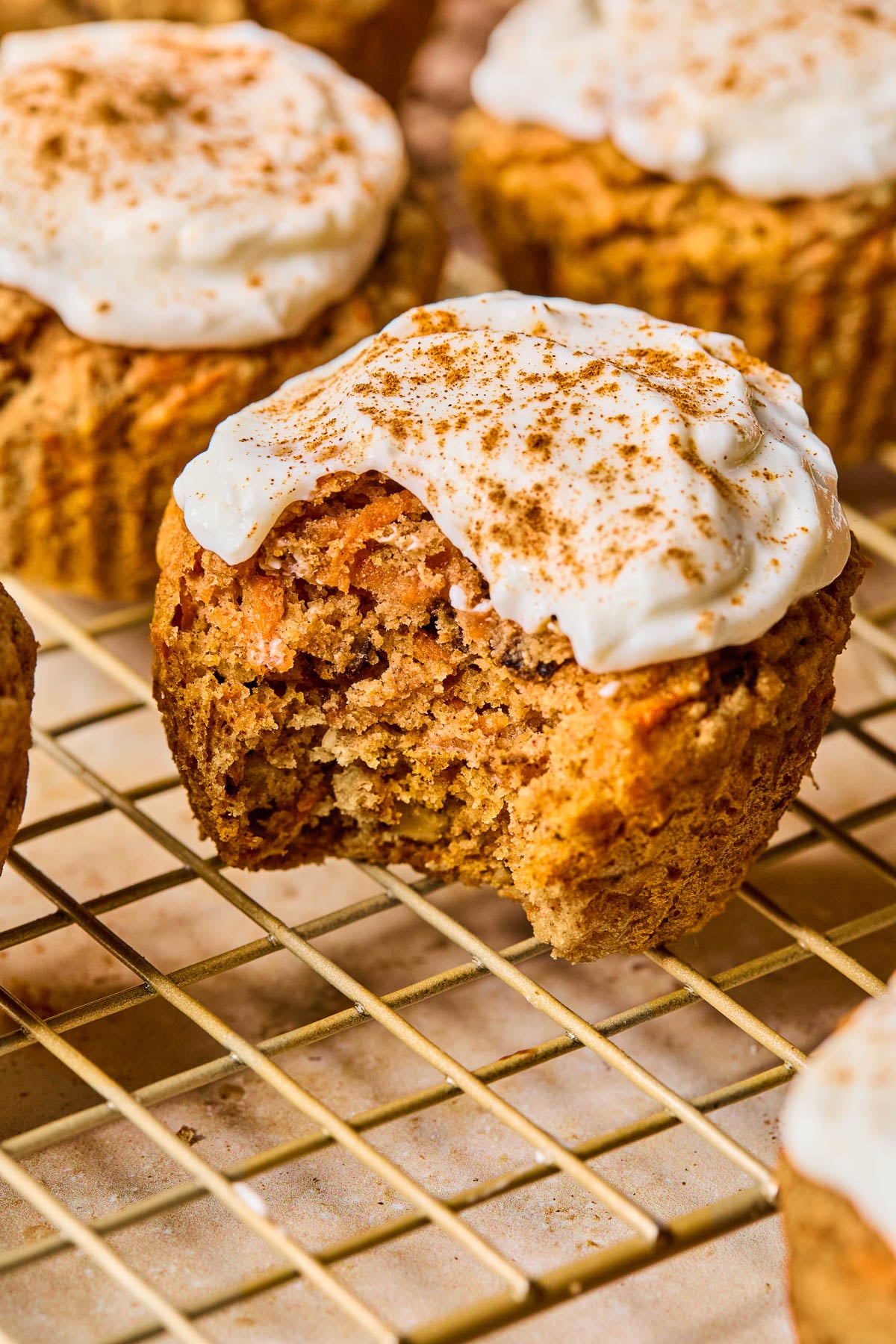 Healthy carrot cake muffins with greek yogurt frosting on a wire cooling rack. The one in front has a bite taken out of it.
