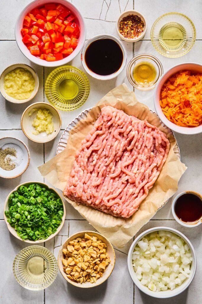 A small bowl of red pepper flakes, a small bowl of fish sauce, a small bowl of honey, a bowl of shredded carrots, a plate of a pound of raw ground chicken, a small bowl of sesame oil, a bowl of diced onions, a bowl of chopped peanuts, a bowl of rice vinegar, a bowl of chopped scallions, a small bowl of salt and pepper, a bowl of minced garlic, a bowl of grated ginger, a bowl of olive oil, a bowl of coconut amino, and a bowl of diced red bell peppers on a grey tile counter.