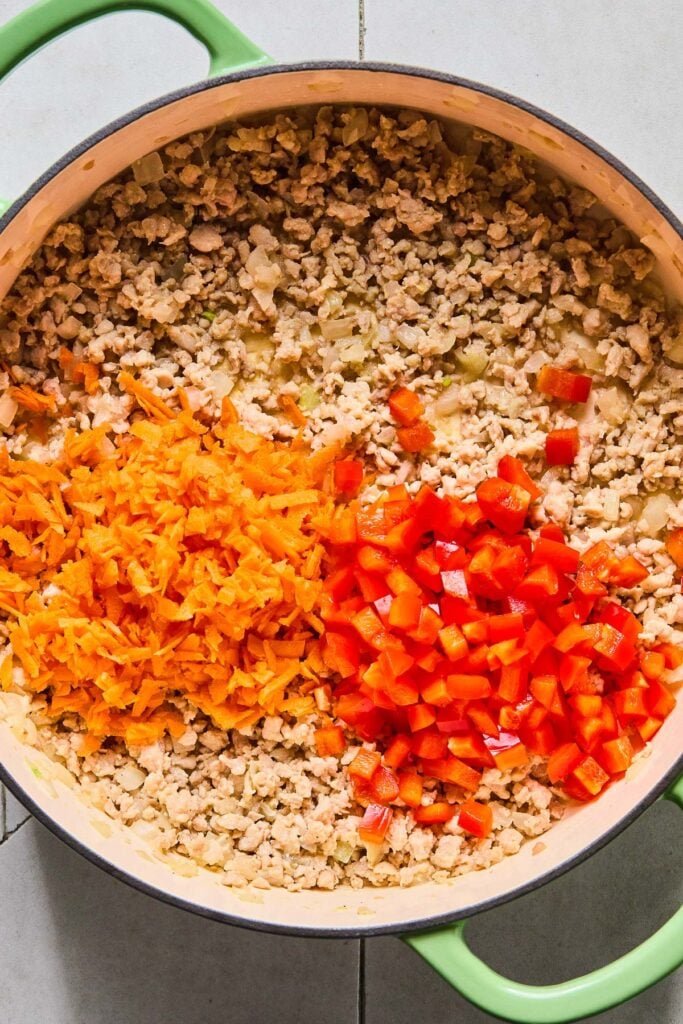 Overhead shot of a large enameled pan filled with saut&eacute;ed ground chicken with a pile of raw shredded carrots and a pile of diced red bell pepper on top.