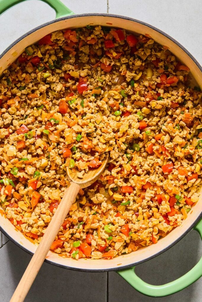 Overhead shot of a large enameled pan filled with saut&eacute;ed ground chicken, onions, red bell peppers, and shredded carrots, green onions, and peanuts all mixed with a stir fry sauce. A wooden spoon is in the pan as well.