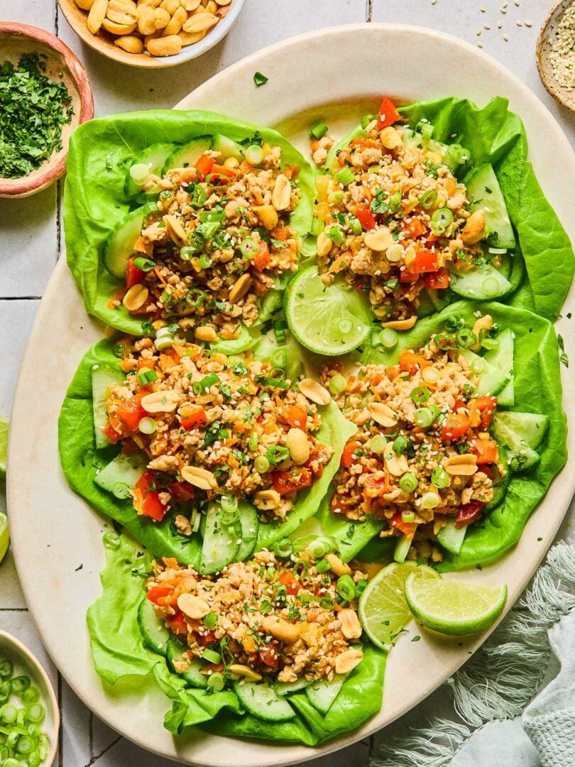 A plate of Asian chicken lettuce wraps with ground chicken, veggies, scallions, peanuts, and sesame seeds. Also on the plate are some lime wedges. Around the plate green kitchen towel, a bowl of sliced green onions, a bowl of cilantro, and a bowl of peanuts.