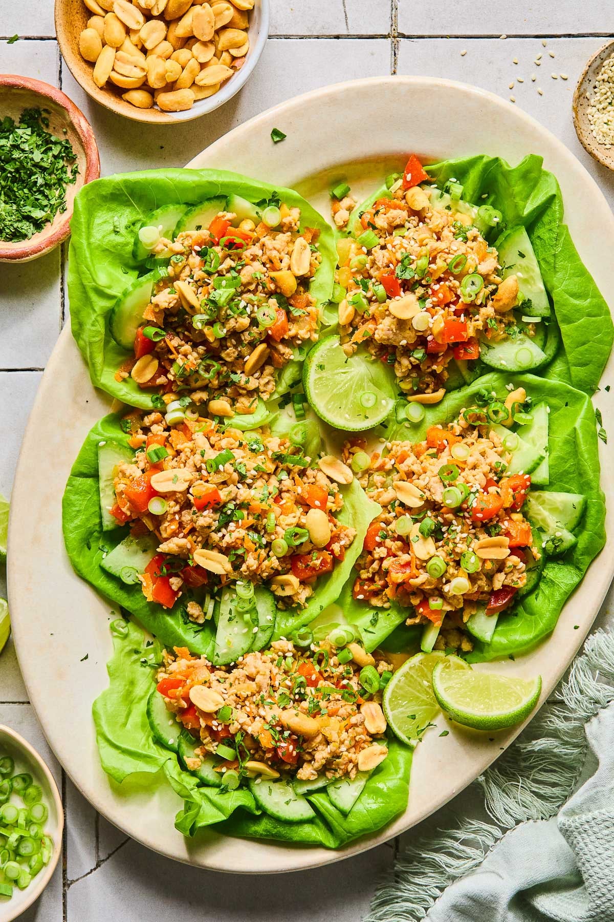 A plate of Asian chicken lettuce wraps with ground chicken, veggies, scallions, peanuts, and sesame seeds. Also on the plate are some lime wedges. Around the plate green kitchen towel, a bowl of sliced green onions, a bowl of cilantro, and a bowl of peanuts.