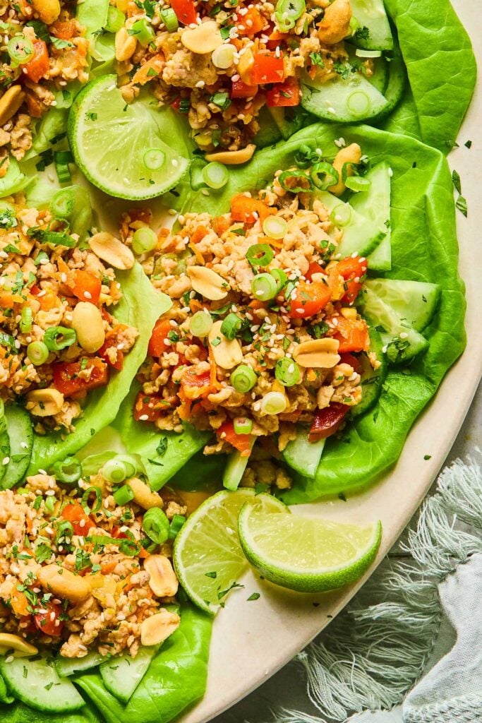 Chicken lettuce wraps with veggies, scallions, peanuts, and sesame seeds on a plate. There are some lime wedges on the plate as well.