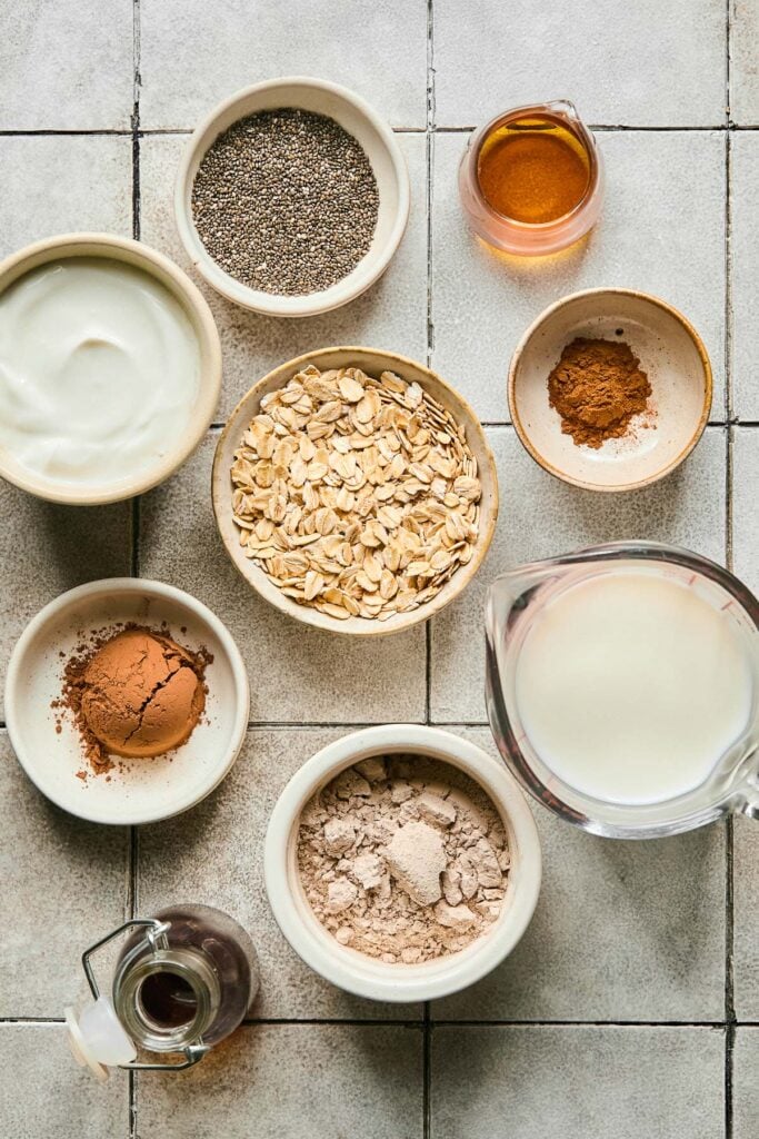 Grey tile countertop with a small jar of maple syrup, a small bowl of cinnamon, a bowl of rolled oats, a cup of milk, a bowl of chocolate protein powder, a jar of vanilla extract, a bowl of cocoa powder, a bowl of greek yogurt, and a bowl of chia seeds on it.