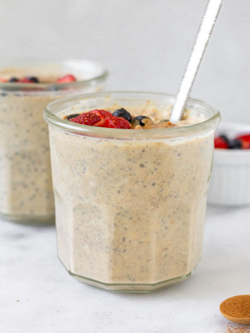 Two jars of overnight oats with protein powder, with one being behind the other. They are topped with berries and a spoon is in the jar in the front. In front of the jar is a teaspoon of cinnamon and off to the back is a dish of berries.