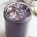 Healthy smoothie in a jar topped with coconut shreds and cacao nibs with a straw. Behind it is a bowl of frozen blueberries and a spoon with peanut butter on it.