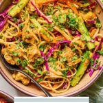 A bowl of noodle salad with chicken, veggies, fresh herbs, and rice noodles in a creamy almond butter sauce. It is topped with sesame seeds, sliced green onions, and more fresh herbs. Around the bowl is a green kitchen towel, another bowl of noodle salad, a jar of dressing, a small bowl of sesame seeds, and a bowl of sliced green onions.
