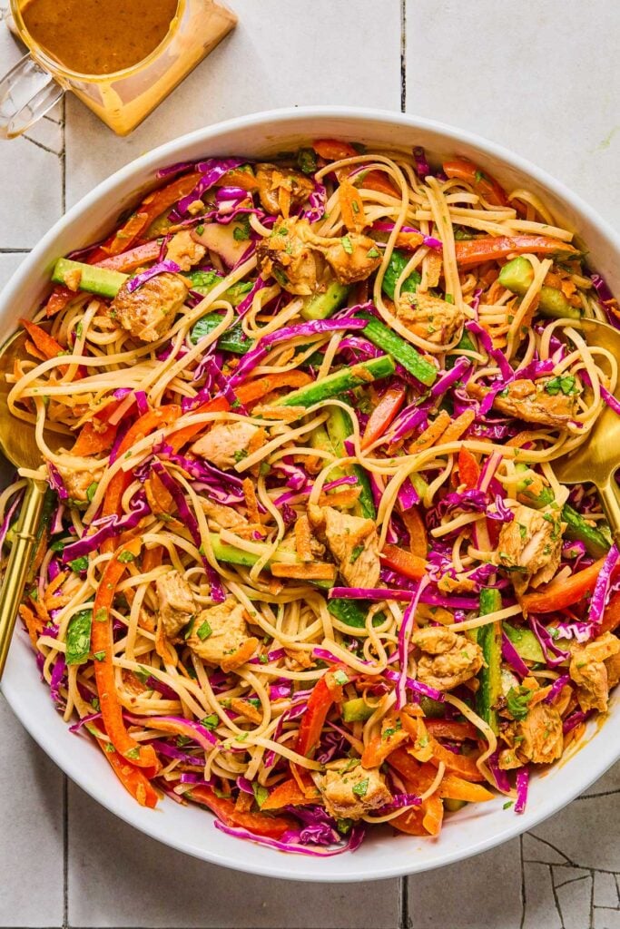 A large bowl of spring roll salad with rice noodles, seasoned chicken, cabbage, cucumber, red bell pepper, carrots, and fresh herbs tossed in a creamy almond butter sauce. Next to the bowl is a jar of more dressing