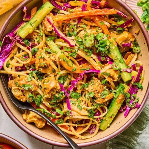 A bowl of noodle salad with chicken, veggies, fresh herbs, and rice noodles in a creamy almond butter sauce. It is topped with sesame seeds, sliced green onions, and more fresh herbs. Around the bowl is a green kitchen towel, another bowl of noodle salad, a jar of dressing, a small bowl of sesame seeds, and a bowl of sliced green onions.