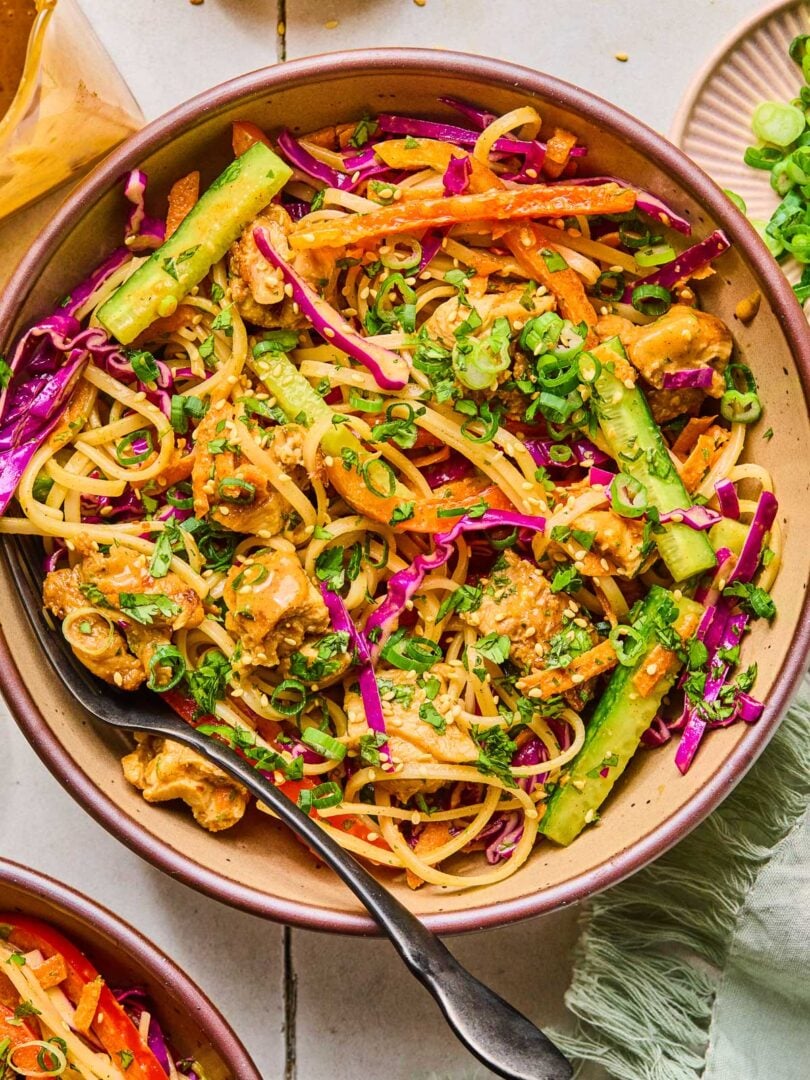 A bowl of noodle salad with chicken, veggies, fresh herbs, and rice noodles in a creamy almond butter sauce. It is topped with sesame seeds, sliced green onions, and more fresh herbs. Around the bowl is a green kitchen towel, another bowl of noodle salad, a jar of dressing, a small bowl of sesame seeds, and a bowl of sliced green onions.