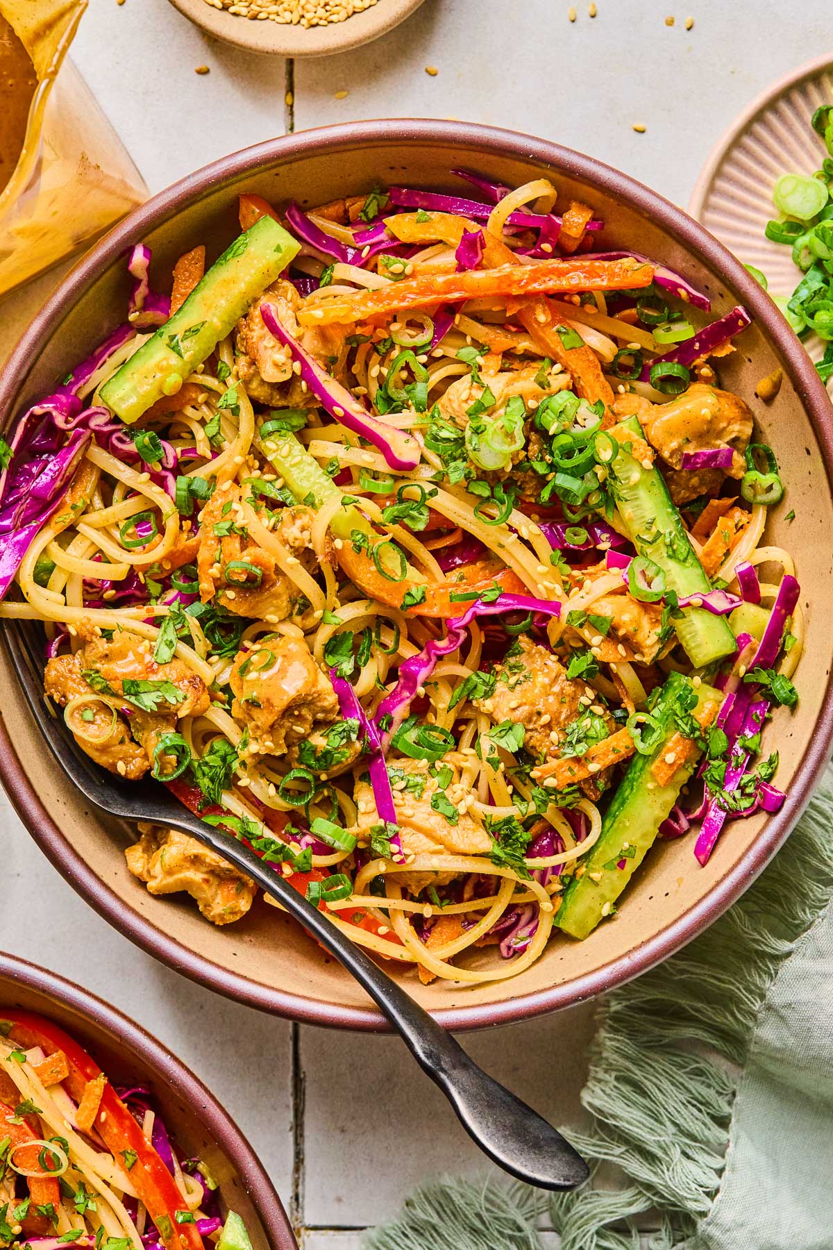 A bowl of noodle salad with chicken, veggies, fresh herbs, and rice noodles in a creamy almond butter sauce. It is topped with sesame seeds, sliced green onions, and more fresh herbs. Around the bowl is a green kitchen towel, another bowl of noodle salad, a jar of dressing, a small bowl of sesame seeds, and a bowl of sliced green onions.