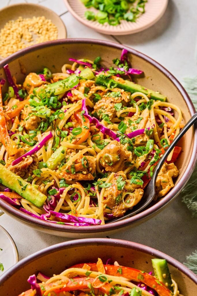 A bowl of Asian noodle salad with rice noodles, fresh veggies, seasoned chicken, and fresh herbs in a creamy sauce. It is topped with sesame seeds, scallions, and fresh herbs. A fork is also in the bowl. Around the bowl is another bowl of Asian noodles salad, a small plate of sesame seeds, and a small plate of sliced scallions.