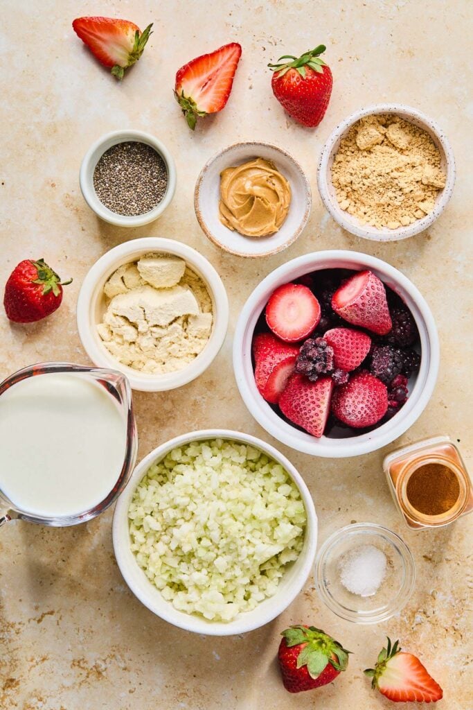 A bowl of peanut butter powder, a small bowl of creamy peanut butter, a bowl of frozen berries, a jar of cinnamon, a small bowl of salt, a bowl of frozen cauliflower rice, a measuring cup of milk, a bowl of vanilla protein powder, and a small bowl of chia seeds on a tan counter. Around the bowls of ingredients are fresh halved strawberries.
