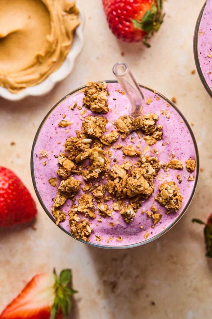 Overhead shot of a peanut butter jelly smoothie in a glass. It is topped with granola, and a straw is in the glass. Around the glass are some fresh strawberries and a bowl of creamy peanut butter.