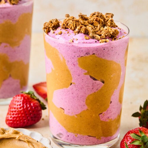 Peanut butter and jelly smoothie in a glass topped with granola. The glass has some peanut butter swirled inside it. Around the glass are some fresh strawberries, a bowl of peanut butter, and another glass with a smoothie in it.