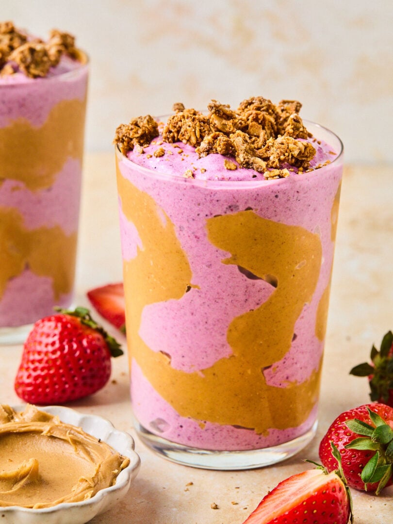 Peanut butter and jelly smoothie in a glass topped with granola. The glass has some peanut butter swirled inside it. Around the glass are some fresh strawberries, a bowl of peanut butter, and another glass with a smoothie in it.