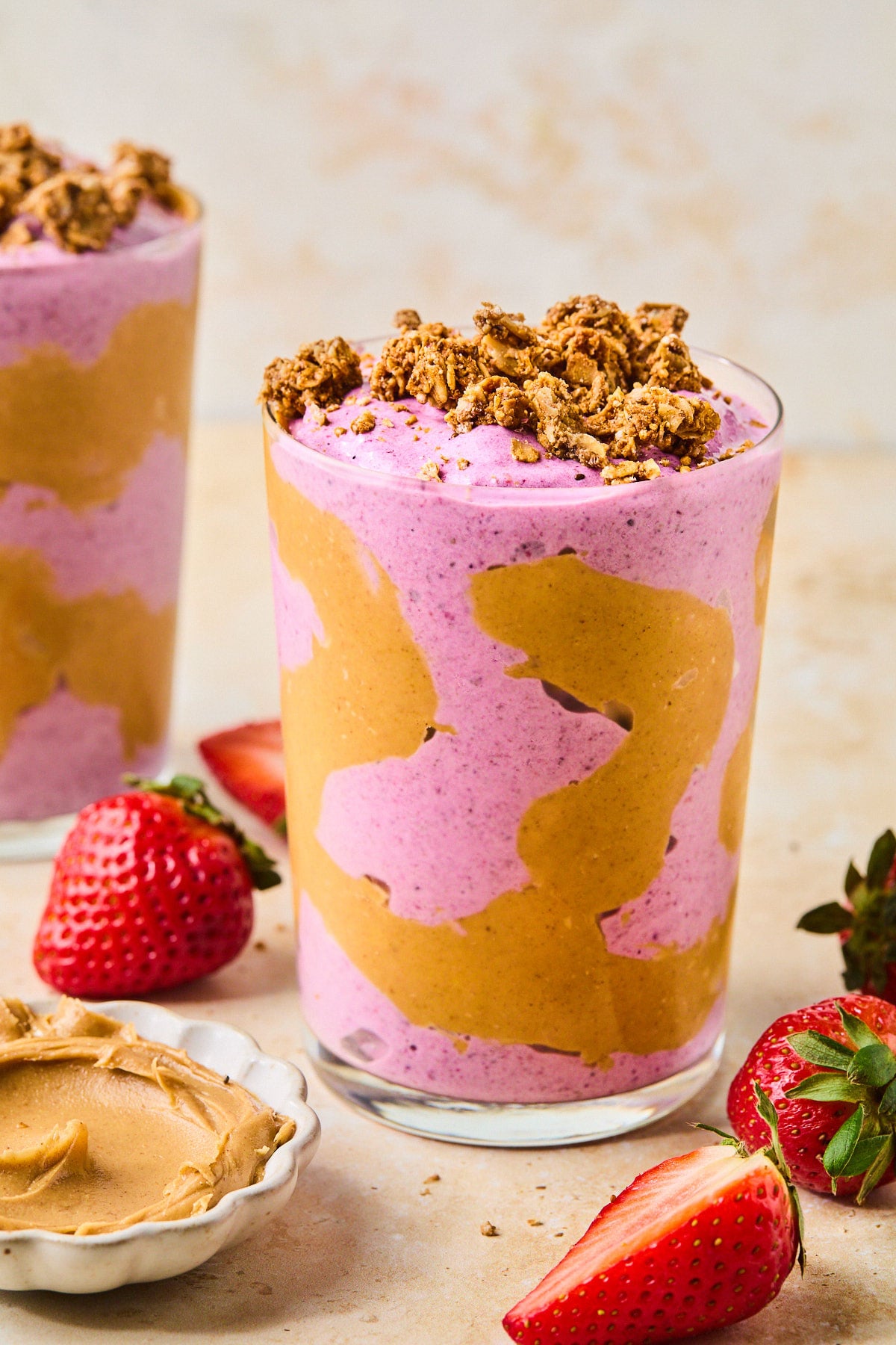Peanut butter and jelly smoothie in a glass topped with granola. The glass has some peanut butter swirled inside it. Around the glass are some fresh strawberries, a bowl of peanut butter, and another glass with a smoothie in it.
