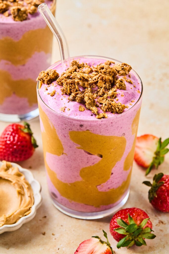 Pb and J smoothie in a glass cup swirled with peanut butter. There is a straw in the cup, and the smoothie is topped with granola. Around the cup are some fresh strawberries, a small bowl of creamy peanut butter, and another smoothie.