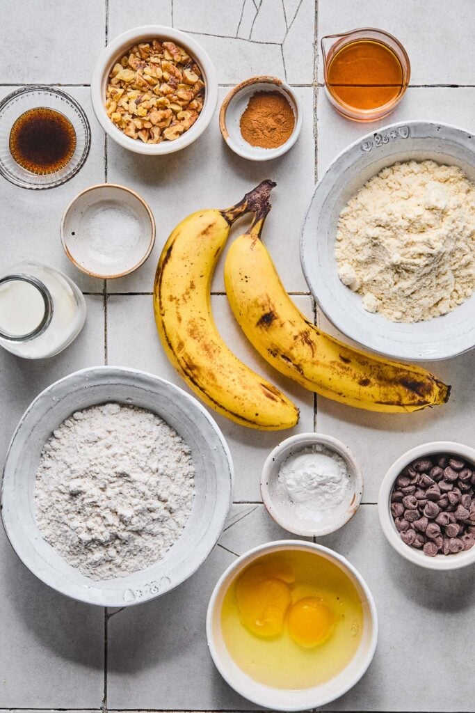 A small measuring cup of maple syrup, a bowl of vanilla protein powder, two spotted bananas, a small bowl of baking powder, a small bowl of chocolate chips, a bowl with 2 cracked eggs in it, a bowl of gluten-free flour, a jar of milk, a small bowl of salt, a small bowl of vanilla bean paste, a small bowl of chopped walnuts, and a small bowl of cinnamon on a white tile counter.