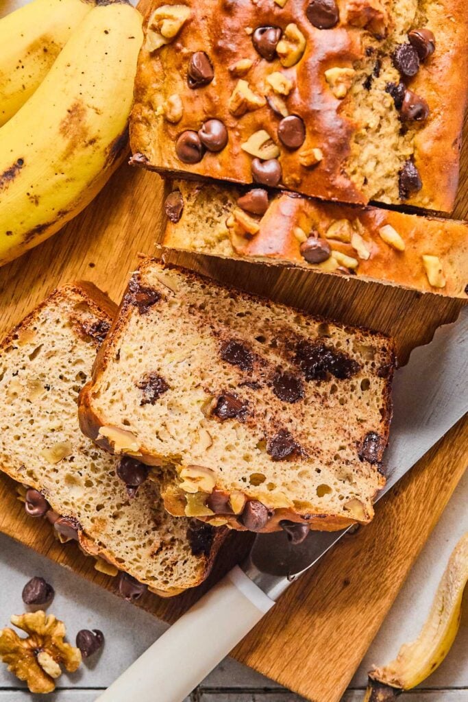 Overhead shot of a loaf of protein banana bread on a cutting board. A few slices have been cut from it. Around the load are some bananas and a knife.