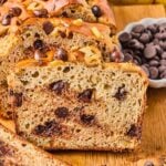 A loaf of protein banana bread with chocolate chips on a wooden cutting board. A few slices have been cut off and are leaning against the loaf. Around the loaf is a bowl of chocolate chips, some chocolate chips and walnuts, a slice of banana bread, and some bananas.