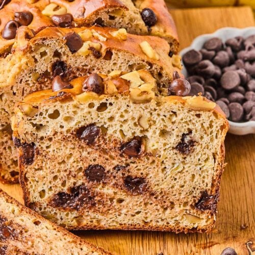A loaf of protein banana bread with chocolate chips on a wooden cutting board. A few slices have been cut off and are leaning against the loaf. Around the loaf is a bowl of chocolate chips, some chocolate chips and walnuts, a slice of banana bread, and some bananas.