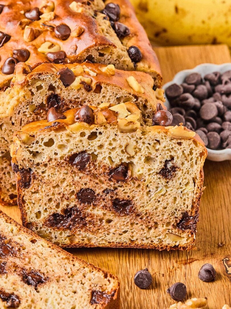 A loaf of protein banana bread with chocolate chips on a wooden cutting board. A few slices have been cut off and are leaning against the loaf. Around the loaf is a bowl of chocolate chips, some chocolate chips and walnuts, a slice of banana bread, and some bananas.