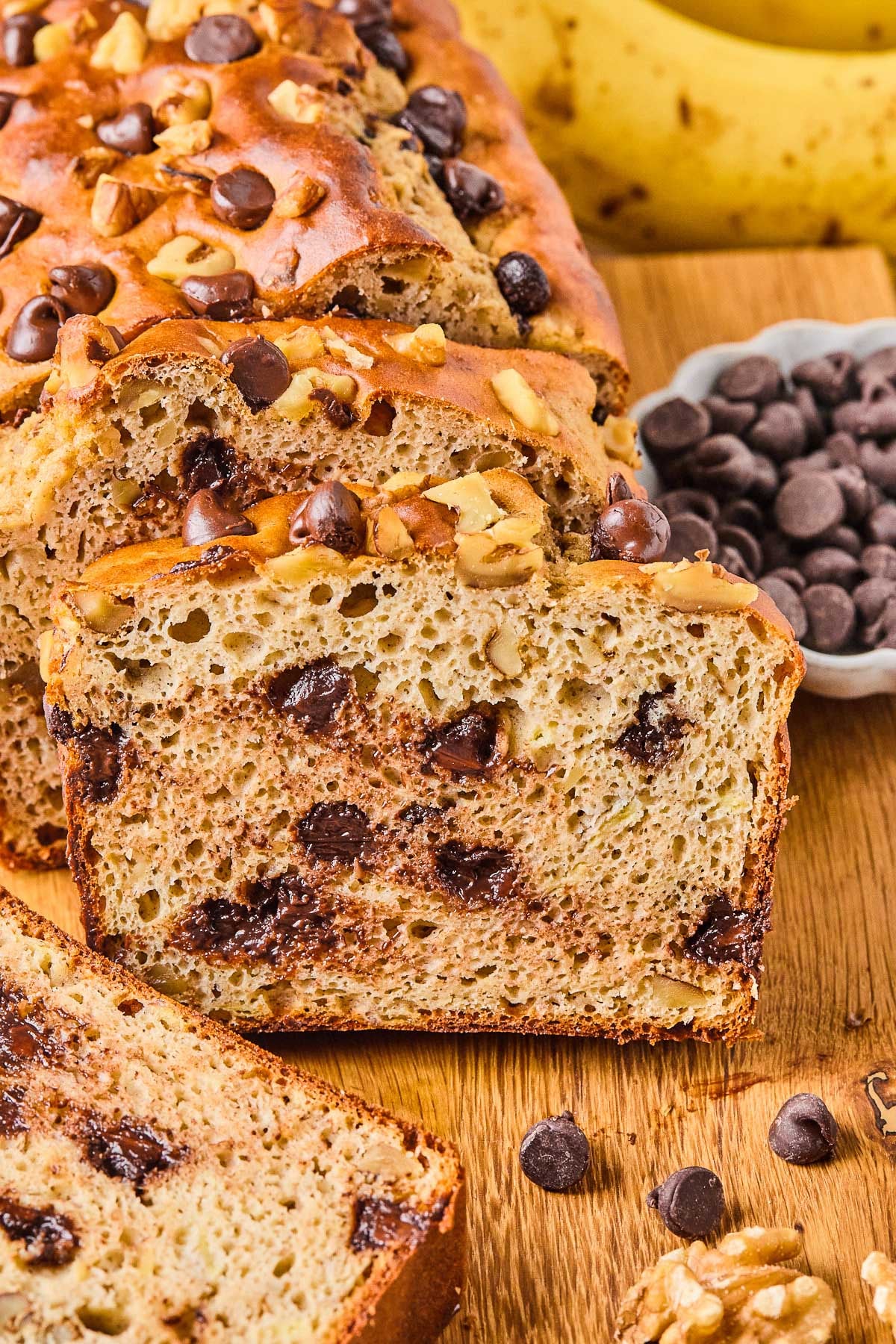 A loaf of protein banana bread with chocolate chips on a wooden cutting board. A few slices have been cut off and are leaning against the loaf. Around the loaf is a bowl of chocolate chips, some chocolate chips and walnuts, a slice of banana bread, and some bananas.
