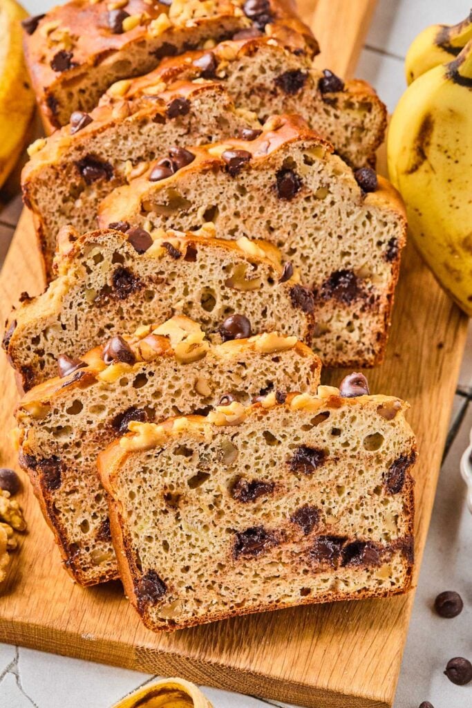 Slices of protein powder banana bread with chocolate chips that have just been cut from the loaf. They are leaning against each other on a wooden cutting board. Around the cutting board are some chocolate chips and bananas.