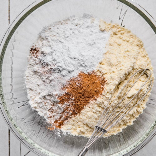 Large mixing bowl of flour, protein powder, baking powder, and spices in a large mixing bowl before it is mixed. A whisk is in the bowl, too.