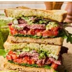 Two halves of a chimichurri steak sandwich with red onion, feta cheese, tomato, and arugula stacked on top of each other on a cutting board. Behind the sandwich is a jar of chimichurri and some wooden salt and pepper shakers.