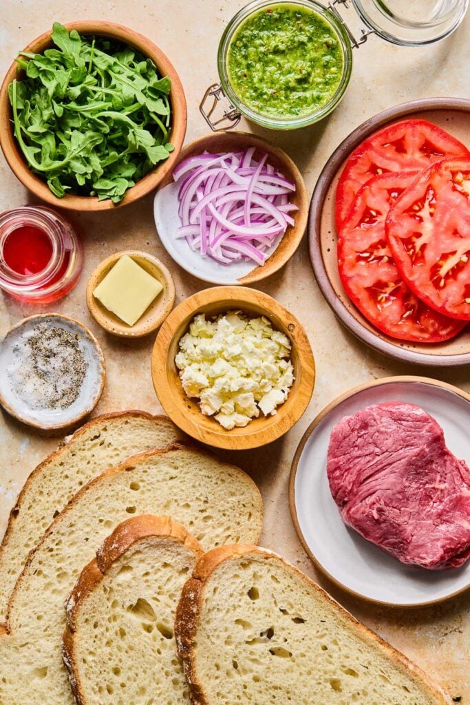 A jar of spicy chimichurri, a plate of sliced tomatoes, a plate with a large piece of raw steak, 4 slices of sourdough bread, a bowl of crumbled feta, a small bowl with a slice of butter in it, a small bowl with salt and pepper, a jar of red wine vinegar, a bowl of sliced red onion, and a bowl of arugula on a tan concrete counter.