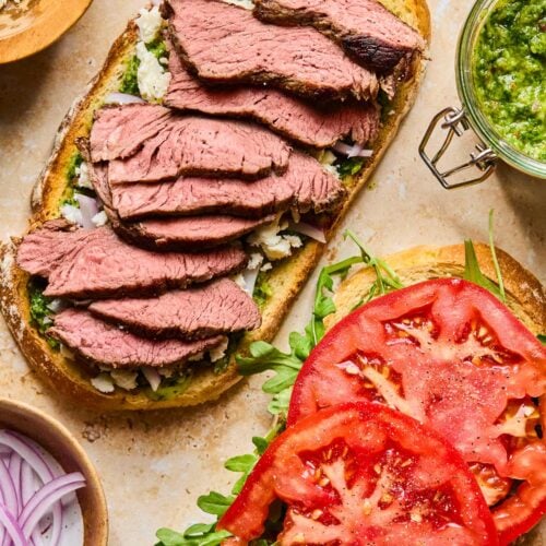 Two pieces of toasted sourdough bread lying on a tan concrete counter. One piece is topped with chimichurri, sliced red onion, crumbled feta, and sliced steak. The other slice is topped with arugula and sliced tomato. Around the bread is a jar of chimichurri, a jar of red wine vinegar, a small bowl of sliced red onion, and a small bowl of feta.