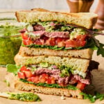 Two halves of a chimichurri steak sandwich with red onion, feta cheese, tomato, and arugula stacked on top of each other on a cutting board. Behind the sandwich is a jar of chimichurri and some wooden salt and pepper shakers.