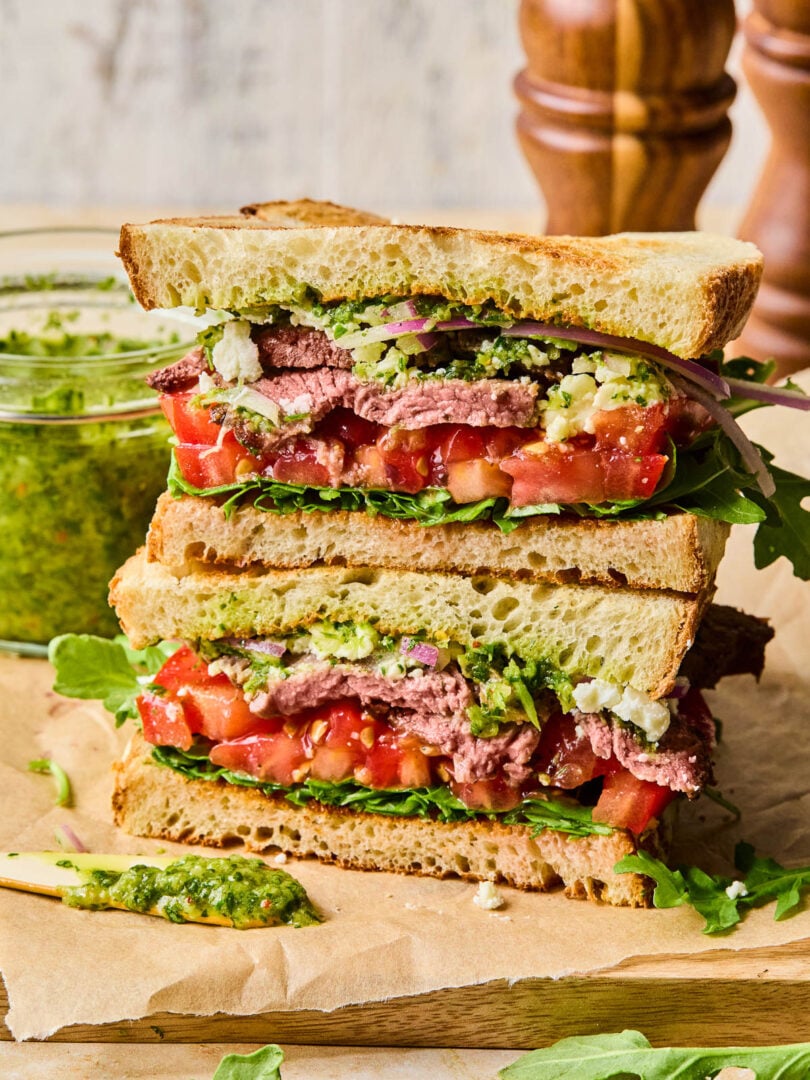 Two halves of a chimichurri steak sandwich with red onion, feta cheese, tomato, and arugula stacked on top of each other on a cutting board. Behind the sandwich is a jar of chimichurri and some wooden salt and pepper shakers.
