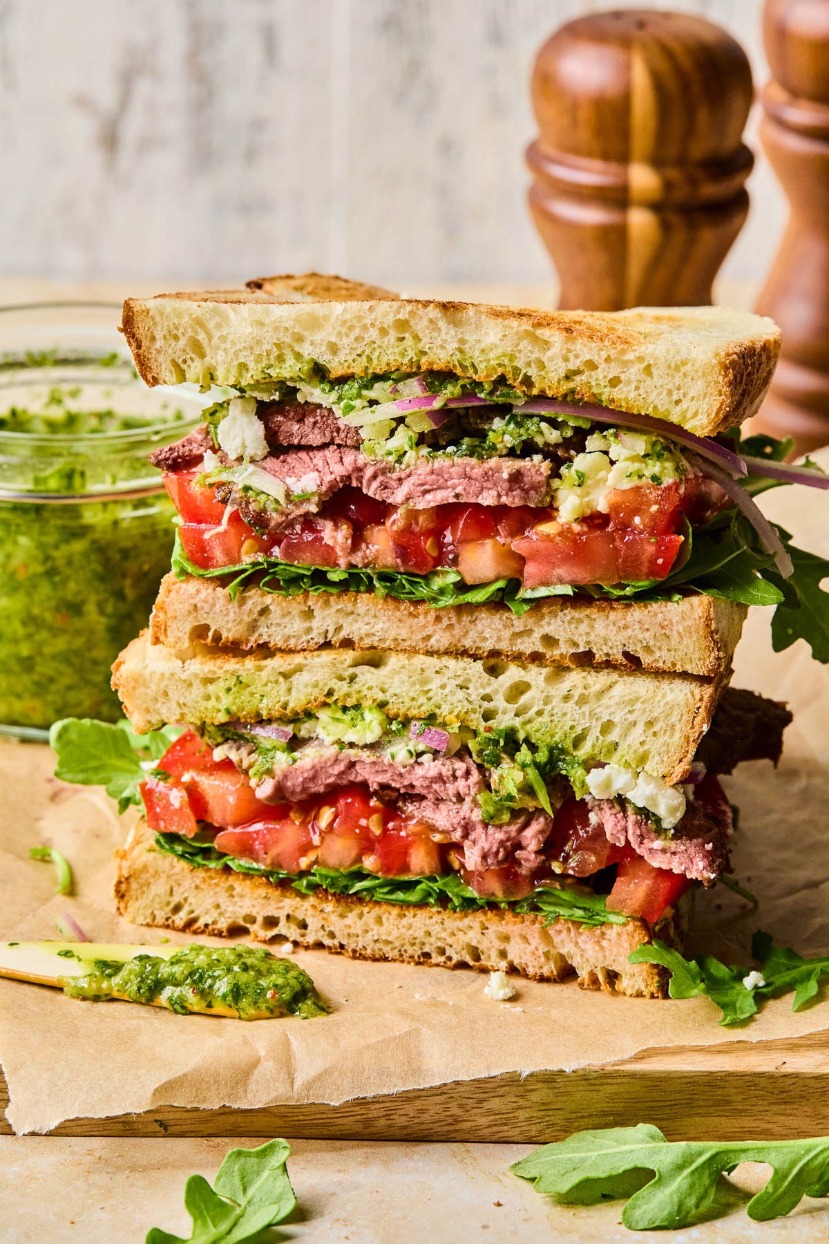 Two halves of a chimichurri steak sandwich with red onion, feta cheese, tomato, and arugula stacked on top of each other on a cutting board. Behind the sandwich is a jar of chimichurri and some wooden salt and pepper shakers.