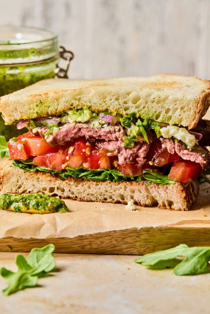 Half of a steak chimichurri sandwich with arugula, feta cheese, tomato, and red onion on a cutting board. Behind the sandwich is a jar of chimichurri sauce.