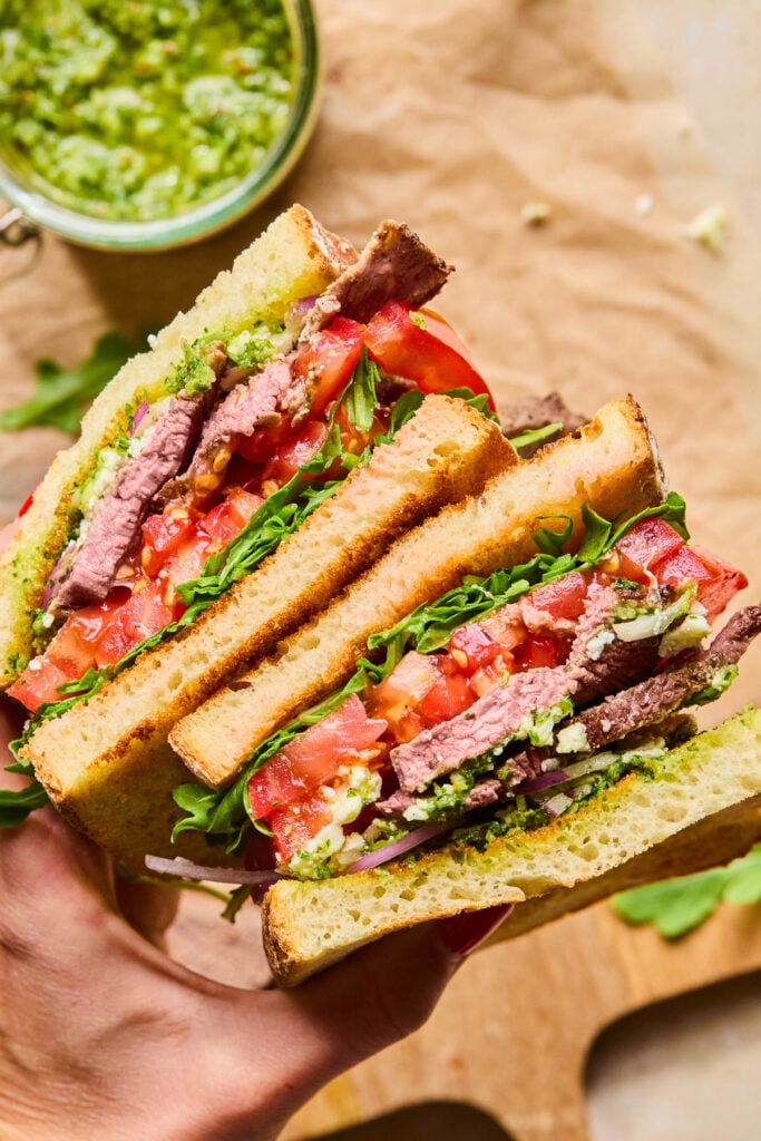 A hand holding up two halves of a steak sandwich with chimichurri, arugula, tomato, red onion, and feta cheese. Underneath the sandwich is a jar of chimichurri sauce on top of some parchment paper on a cutting board.