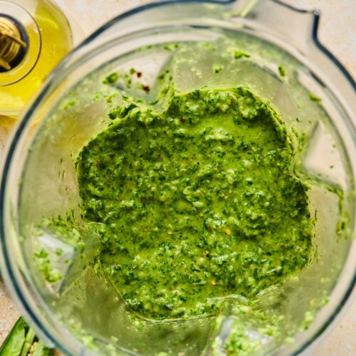 Blender chimichurri sauce in a blender. Around the blender is half a jalape&ntilde;o, a jar of olive oil, and a juiced lime.