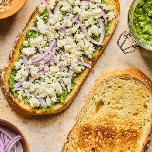 Two pieces of toasted sourdough bread lying on a tan concrete counter. One piece is topped with chimichurri, sliced red onion, and crumbled feta. Around the bread is a jar of chimichurri, a small bowl of sliced red onion, and a small bowl of feta.