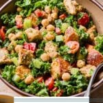 Kale caesar salad with chicken, veggies, and croutons in a bowl with a fork in it. Around the bowl is a small bowl of croutons, another bowl of salad, and a glass with dressing in it.