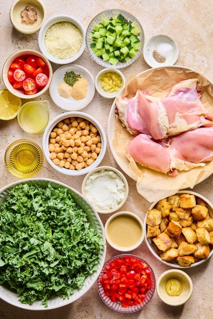 A bowl of diced cucumbers, a small bowl of salt and pepper, a small bowl of minced garlic, a plate of raw chicken thighs, a bowl of sourdough croutons, a small bowl of dijon mustard, a bowl of diced red bell peppers, a bowl of tahini, a bowl of greek yogurt, a large bowl of kale, a cup of olive oil, a bowl of chickpeas, a small pitcher of lemon juice, half a lemon, a small plate with a few piles of spices on it, a bowl of quartered cherry tomatoes, a bowl of grated parmesan cheese, and a small bowl of anchovy paste on a tan concrete counter.