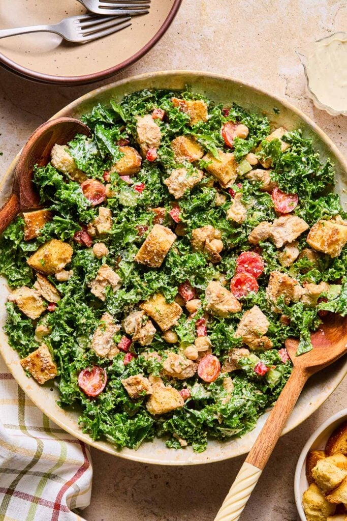 Caesar kale salad with chicken, veggies, croutons, and cheese in a large bowl with salad servers in it. Around the bowl are a checkered kitchen towel, a bowl with two forks, a pitcher with caesar dressing, and a bowl of croutons.