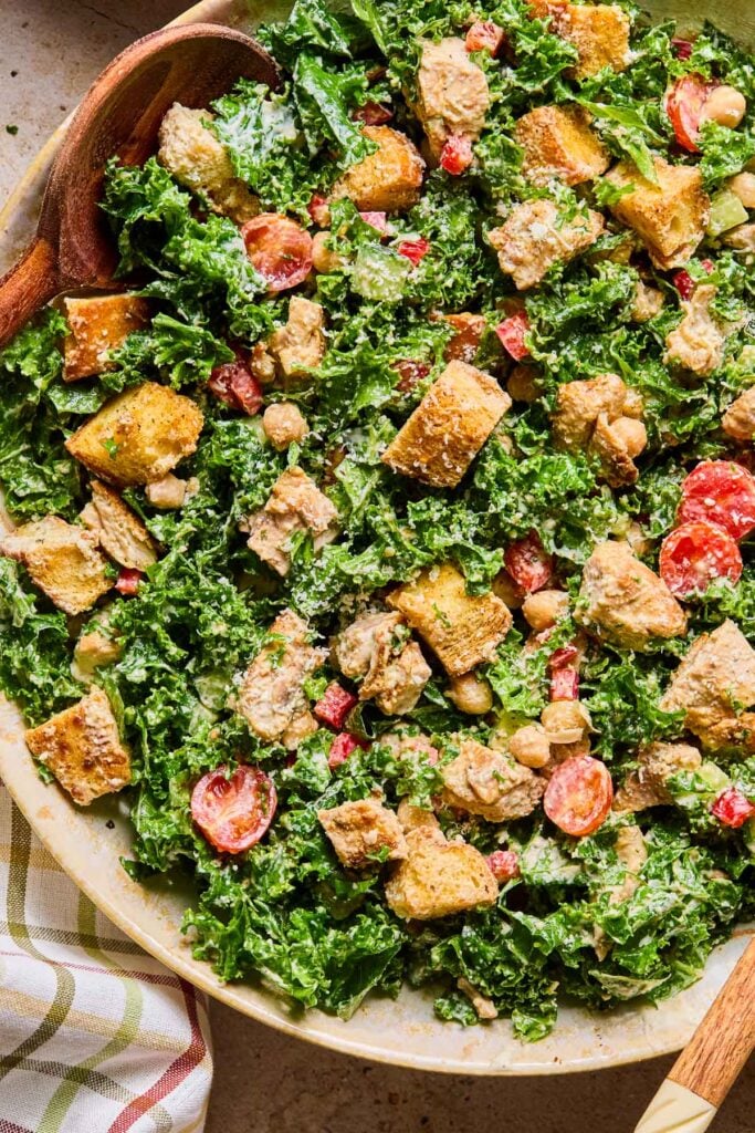 A large bowl of caesar kale salad with chicken, veggies, and croutons with wooden salad servers. Next to the bowl is a checkered kitchen towel.