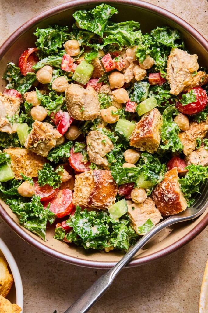 Overhead shot of a kale chicken caesar salad with veggies, croutons, and parmesan cheese in a bowl with a fork in it.