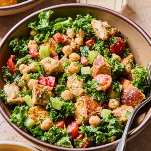Kale caesar salad with chicken, veggies, and croutons in a bowl with a fork in it. Around the bowl is a small bowl of croutons, another bowl of salad, and a glass with dressing in it.