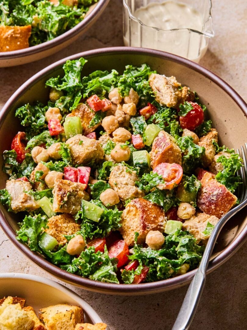 Kale caesar salad with chicken, veggies, and croutons in a bowl with a fork in it. Around the bowl is a small bowl of croutons, another bowl of salad, and a glass with dressing in it.