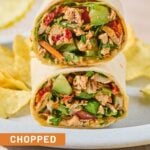 Two halves of a chicken salad wrap, with veggies stacked on top of each other on a plate, along with potato chips.