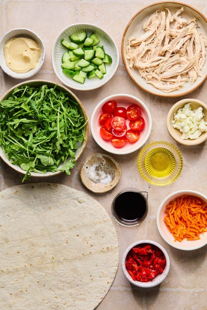 A bowl of shredded chicken, a small bowl of crumbled goat cheese, a small bowl of olive oil, a small bowl of shredded carrots, a small bowl of balsamic vinegar, a large burrito size tortilla, a small bowl of salt and pepper, a bowl of arugula, a bowl of halved cherry tomatoes, a bowl of diced cucumbers, and a bowl of hummus on a tan tile counter.